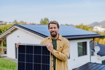 Mature man holding solar panel near his house with solar panels on the roof. Alternative energy,...