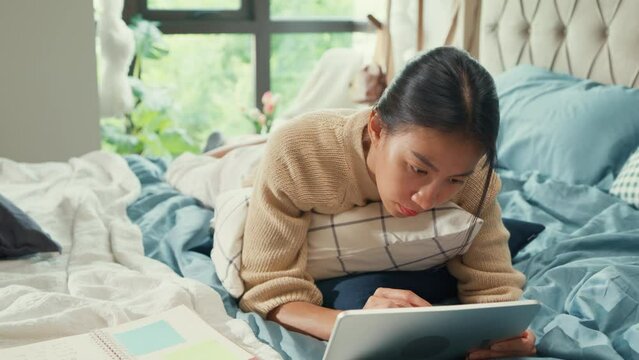 Happy Asian girl university with white cream pajamas lay down on fluffy comfort bed use digital tablet learning online in bedroom at home. Revisualizing flexible working, Work from home concept.