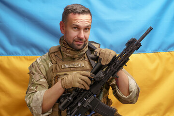 Soldier of Ukrainian Army handsome man holding rifle in a camouflage ammunition posing on Ukrainian flag background. Brave Ukrainian army man with weapon. The flag of Ukraine on background