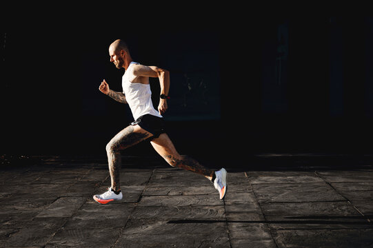 Bald young athletic tattooed man running fast outdoor, sportsman preparing to marathon, doing cardio exercises on the open air, side view