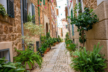 Mediterranean alley in the old town of Mallorca with many green plants in flower pots. Cobblestones lead between beige brick houses. Colorful flags are stretched between them.
