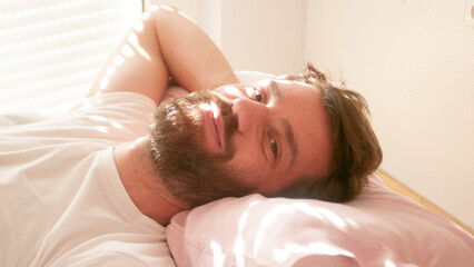 Hombre blanco barbudo tumbado en la cama al sol mirando a cámara