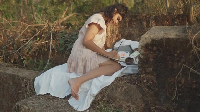 Slow motion handheld shot of a young pretty artist from india sitting on a white linen cloth on stones and painting an abstract painting in her garden