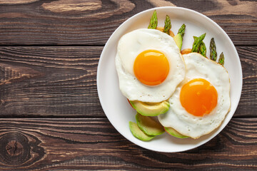 Tasty breakfast. Fried eggs with green asparagus and avocado on bread toasts on the plate on wooden table. Top view