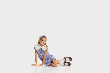 Beautiful little cute stylish girl, kid in retro fashion outfit sitting on floor and looking at camera isolated on white background. Concept of kids emotions, facial expression, beauty.