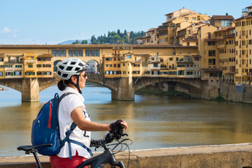 cheerful senior woman cycling with her electric mountain bike in downtown of Florence  with famous bridge of ponte Vecchio in background, Tuscany, Italy