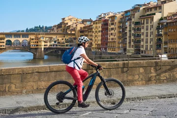 Gartenposter cheerful senior woman cycling with her electric mountain bike in downtown of Florence  with famous bridge of ponte Vecchio in background, Tuscany, Italy © Uwe