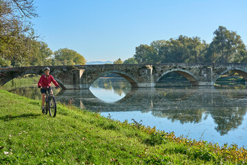 nice senior woman riding her electric mountain 
at river Arno near Arezzo, with famous bridge of Ponte Buriano in background, Arezzo,T uscany , Italy