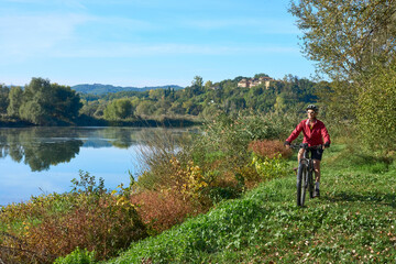 Fototapeta premium nice senior woman riding her electric mountain at river Arno near Arezzo, with famous bridge of Ponte Buriano in background, Arezzo,T uscany , Italy
