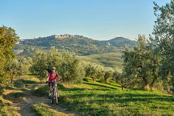 Obraz premium nice senior woman riding her electric mountain bike between olive trees in the Ghianti area with medieval city of Montepulciano in background, Tuscany , Italy
