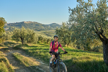 Fototapeta premium nice senior woman riding her electric mountain bike between olive trees in the Ghianti area with medieval city of Montepulciano in background, Tuscany , Italy