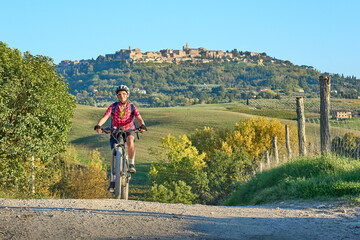 nice senior woman riding her electric mountain bike between olive trees in the Ghianti area with medieval city of Montepulciano in background, Tuscany , Italy