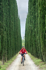 Obraz premium nice senior woman riding her electric mountain bike in a cypress avenue in the Chianti area near Pienza, Tuscany , Italy