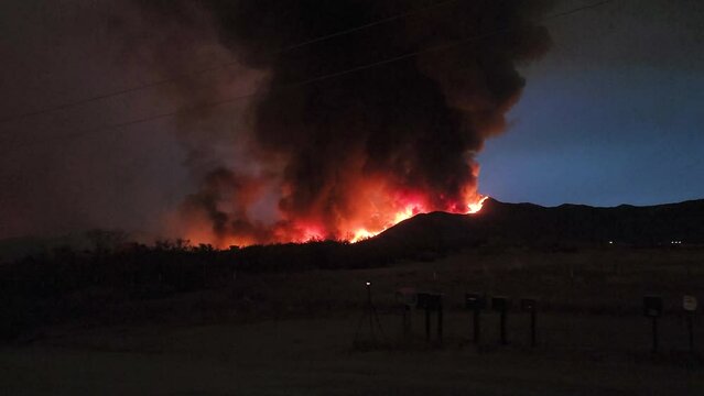 night time wildfire wide establishing shot of flames burning mountain side with dark black smoke plumes rising into the sky at night with mailboxes in the foreground showing rural devastation 
