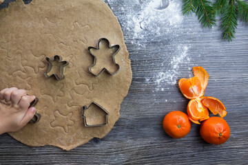 New Year and Christmas decorations on a wooden surface sprinkled with flour with tangerines and a Christmas tree. The hands of a small child make molds for cooking gingerbread in the form of a man.