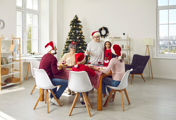 Family celebrating Christmas. Older family member makes toast during traditional festive family Christmas dinner. Multi-generational family in Santa hats gathered together at festive table at home.