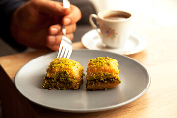 He eats Turkish baklava with green pistachio in plate on an oak coffee table in home environment, with a fork and accompanied by Turkish coffee