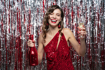Woman in red festive dress holding champagne bottle and glass celebrating new year on tinsel...