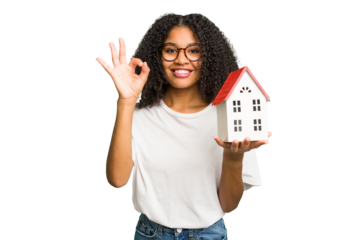 Young african american woman moving to a new home while picking up a box full of things isolated cheerful and confident showing ok gesture.