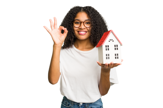 Young african american woman moving to a new home while picking up a box full of things isolated cheerful and confident showing ok gesture.