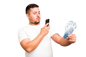 Young caucasian man holding banknotes and mobile phone isolated