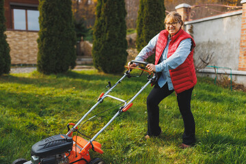 Middle aged woman in red vest using lawn mower on backyard, looking at camera. Female gardener...