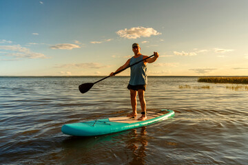 A man in shorts on a SUP board with a paddle floats on the water in the rays of the setting sun.