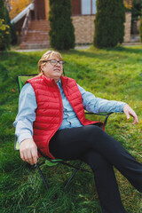 Naklejka na ściany i meble Middle aged woman in red vest relaxing in a chair on a grass meadow outdoor. She is absolutely happy. Slow living, gardening hobby concept