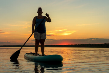 A woman in shorts and a T-shirt with a mohawk on a SUP board in the evening at sunset in the lake.