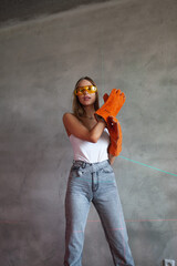 Beautiful blonde girl stands with construction tools in construction uniform in orange gloves and yellow glasses