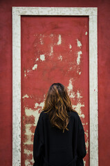 Una donna  con un caotto nero e lunghi caelli castani fotografata di spalle di fronte al muro rosso di di una vecchia casa