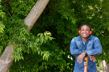 Against backdrop green foliage in city park, photo of smiling dark-skinned man with his hands on the steering wheel of a scooter. Riding an electric scooter, an African listens to music enjoys nature