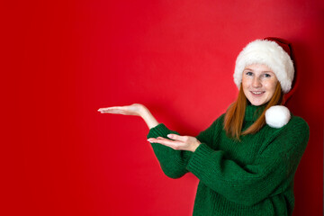 Fototapeta na wymiar Merry Christmas Portrait of a beautiful young teenage girl in a cozy knitted green sweater and Santa's hat. The red background is the place for the text.
