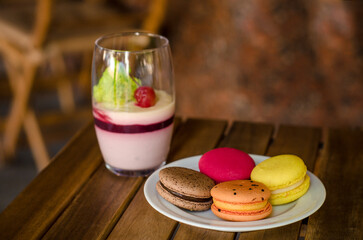 Large transparent glass with cherry dessert, different types of macaron cake on a white saucer standing on the outdoor terrace of cafe, on a wooden table against background of the windows of cafe