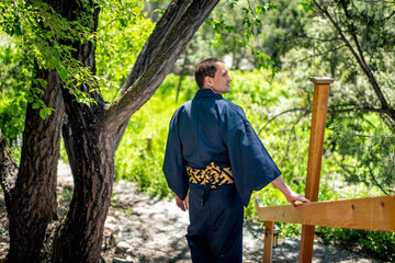 Young happy smiling man in kimono costume walking, leaning on railing fence in outdoor garden in Japan with nature view, looking back turning head