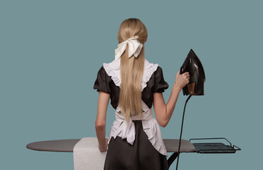 A woman dressed as a maid irons a towel on an ironing board. Back view. Blue background.