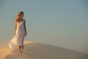 Fototapeta na wymiar A girl in a white dress fluttering in the wind walks through the desert in the sunset light.