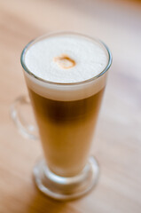 Beautiful tall transparent glass with a handle containing a latte coffee drink on a table, in a cafe in front of a window with natural light