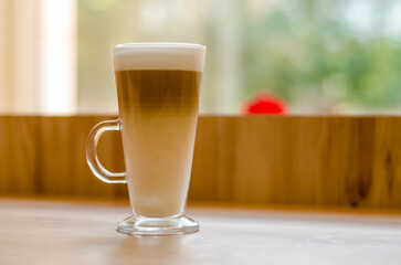 Beautiful tall transparent glass with a handle containing a latte coffee drink on a table, in a cafe in front of a window with natural light