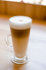 Beautiful tall transparent glass with a handle containing a latte coffee drink on a table, in a cafe in front of a window with natural light