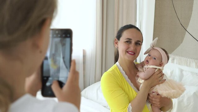Little girl taking photos of her cute baby sister and mother on smartphone in bedroom. Happy life moments and family lifestyle concept