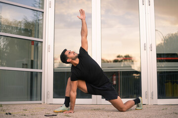 Boy dressed in black workout clothes is performing stretching on the ground.he is training on the street during the autumn season next to some glass windows.
