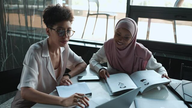 Two happy multi-ethnic Muslim and African women talking and looking at laptop in the office