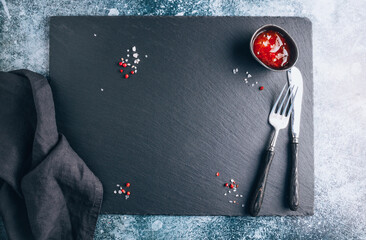 Dark food background with empty black stone board, chili sauce, spicies, knife, fork and napkin. Free space for text or product.