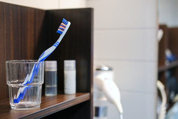 Nahaufnahme einer Zahnbürste in einem Glas auf einem Regal im Badezimmer eines Hotels mit weiteren Pflegeutensilien und Föhn im verschwommenen Hintergrund, selektiver Fokus