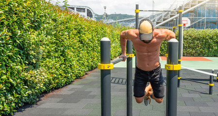 Young man with a naked torso and dark shorts and a cap, trains doing push-ups on the uneven bars on...