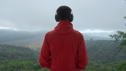 Adult man listening to music through large headphones in mountains on stormy day looking at valley and misty volcano. Music-lover on a hiking trip wearing red jacket enjoying mount view. soft focus.