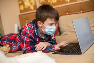 teenage boy in protective medical mask coughs in fist. child remotely does lessons lying on bed near laptop. Online shopping, e-learning, remote studying on internet concept.