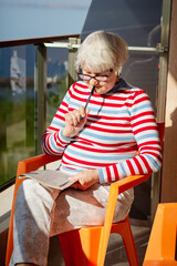 Senior woman in glasses sitting on a balcony near the sea and looking at magazine, solving a crossword