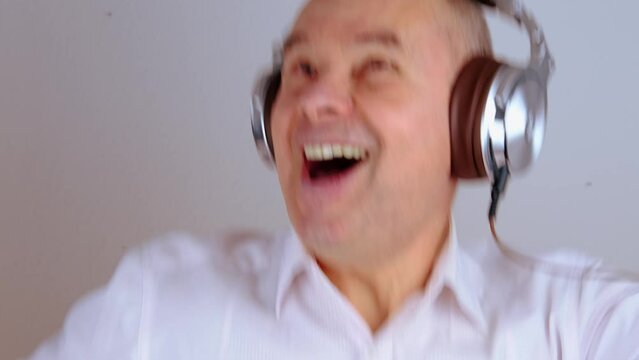 close-up of mature charismatic man 60 years in headphones listening to music with emotions on face gestures, on light gray background, concept learning foreign languages, live streaming and vlogging